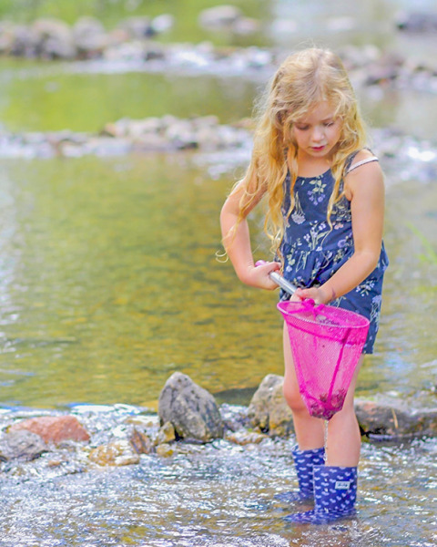 Niña rubia con botas azules explora un arroyo con una red rosa en Camping Paradis Le Giessen en Grand Est, Francia.