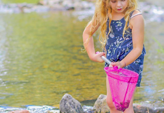 Bambina bionda con stivali blu esplora un ruscello con un retino rosa a Camping Paradis Le Giessen, Grand Est.