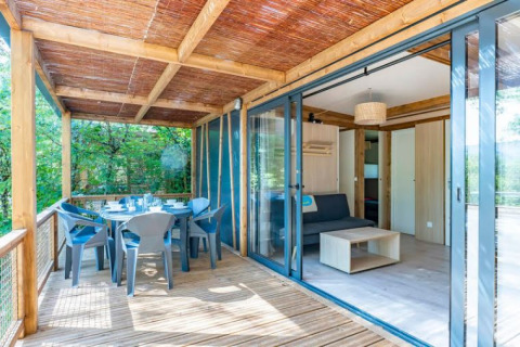 Terraza de madera y sala de una Cottage Premium 3 en Camping Paradis Le Giessen, rodeada de árboles en Francia.