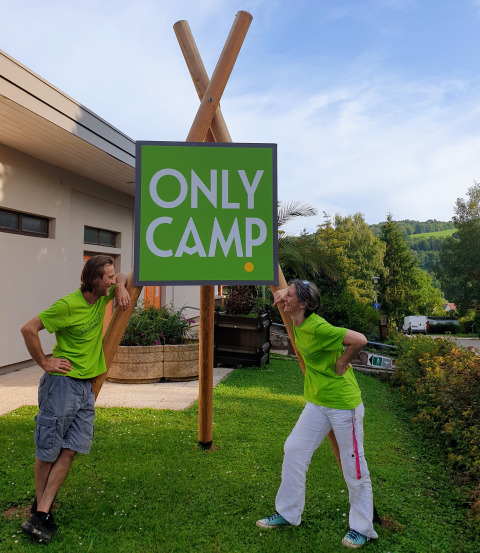 Zwei Personen in grünen Shirts stehen neben einem 'ONLY CAMP'-Schild auf dem Camping l'Orée d'Alsace in Frankreich.