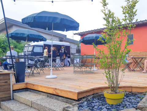 Buitenterras met tafels, parasols, gasten en een foodtruck op Onlycamp Camping l'Orée d'Alsace in Grand Est, Frankrijk.
