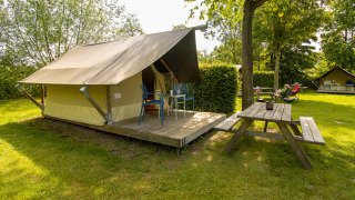 Safari Trekking tent op een groene camping met houten terras, stoelen en picknicktafel in de buurt.