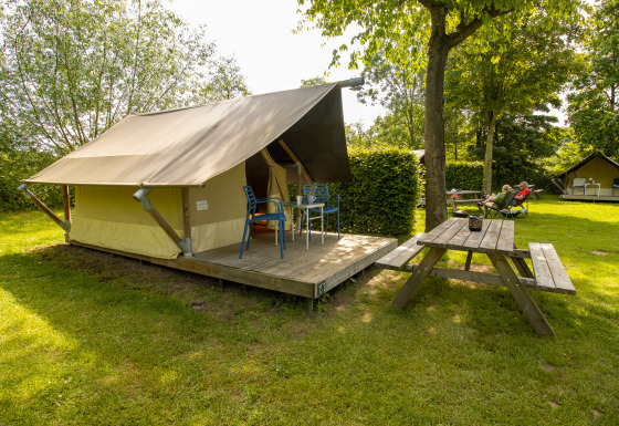 Safari Trekking tent op een groene camping met houten terras, stoelen en picknicktafel in de buurt.