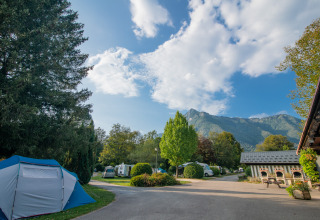 Campingplads med telte og campingvogne omgivet af træer og bjerge under en blå himmel i Auvergne-Rhône-Alpes.