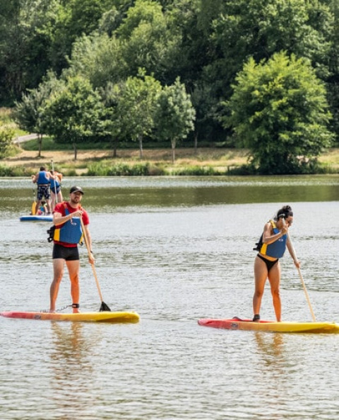 Deux personnes pratiquent le paddle sur un lac à Onlycamp Camping Le Lac de Roybon, entourées de verdure.