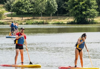 Zwei Personen paddeln auf Paddleboards auf einem See im Onlycamp Camping Le Lac de Roybon, umgeben von Bäumen.