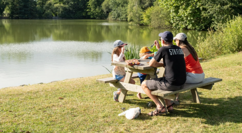 Family enjoys a picnic by the lake at Onlycamp Camping Le Lac de Roybon in the scenic Auvergne-Rhône-Alpes, France.