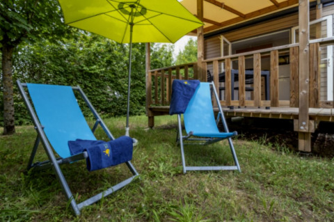 Two blue deck chairs and a yellow umbrella outside a lodge at Seasonova Camping Le Plan d’Eau in France.
