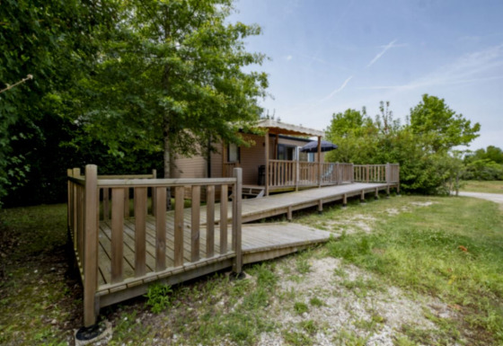 Mobile home Comfort MIVA with wooden deck and ramp, surrounded by trees and greenery in a rural area.