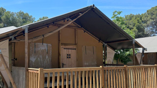 Safari-Zelt namens Nomad mit Holzveranda und Geländer, umgeben von Bäumen unter blauem Himmel.