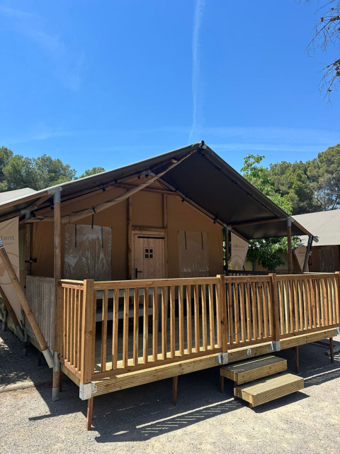 Safari tent named Nomad with wooden porch and railing, set under a clear blue sky and surrounded by trees.