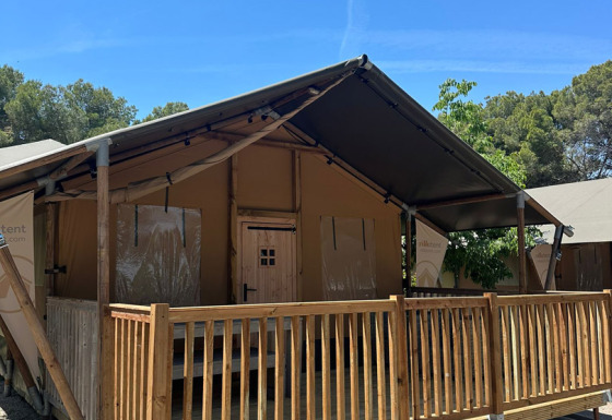 Safari tent named Nomad with wooden porch and railing, set under a clear blue sky and surrounded by trees.