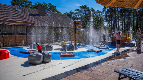Zona de juegos acuáticos junto a un lodge, con fuentes de agua, estructuras y niños jugando al sol.