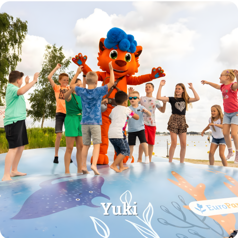 Niños juegan con una gran mascota naranja de pelo azul sobre una colchoneta hinchable cerca de una cabaña.