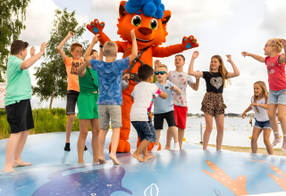 Niños juegan con una gran mascota naranja de pelo azul sobre una colchoneta hinchable cerca de una cabaña.