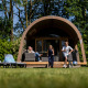 Kinderen spelen voor een grote safari tent lodge XL met sanitair, terwijl een volwassene ontspant op de veranda.