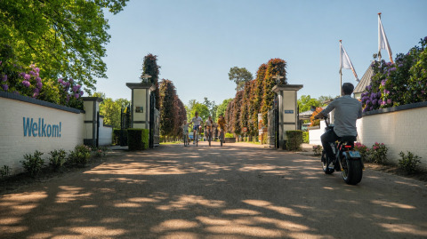Entrée d’un lodge avec des cyclistes et un homme sur un vélo électrique. Panneau 'Welkom!' à gauche.