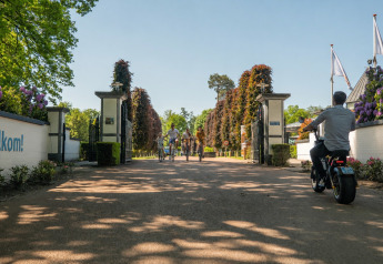 Ingang van een lodge met mensen op fietsen en een man op een e-bike. Links het bord 'Welkom!' op de muur.