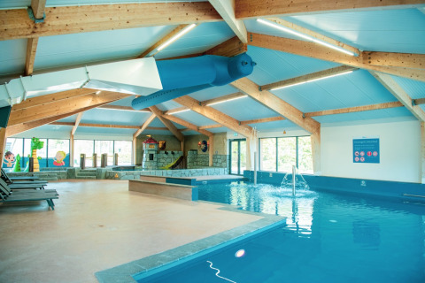 Espace piscine intérieure dans un lodge avec toboggan, chaises longues et coin enfants coloré en arrière-plan.