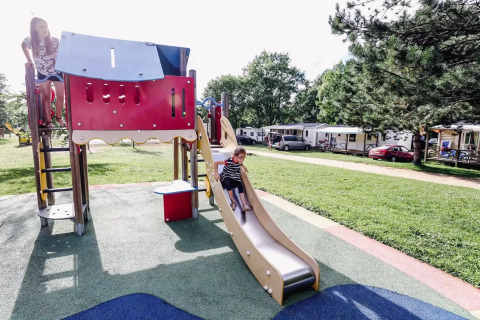 Kinder spielen auf einem Spielplatz bei Seasonova Camping Le Brabois, mit Wohnwagen und Bäumen im Hintergrund.
