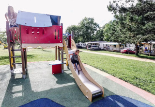 Kinder spielen auf einem Spielplatz bei Seasonova Camping Le Brabois, mit Wohnwagen und Bäumen im Hintergrund.