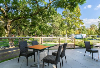 Außenterrasse mit Tischen und Stühlen im Seasonova Camping Le Brabois, Grand Est, Frankreich, von Bäumen umgeben.