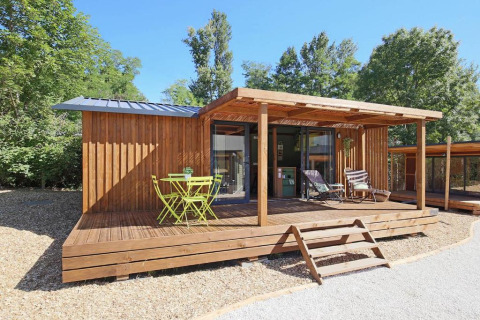 Casa pequeña Premium cottage con terraza de madera, muebles exteriores verdes y rodeada de árboles soleados.