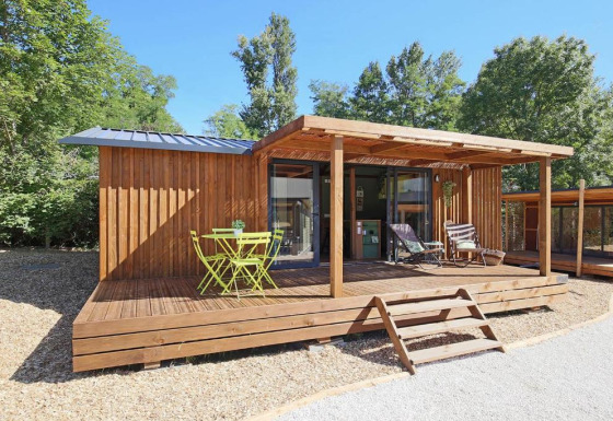 Casa pequeña Premium cottage con terraza de madera, muebles exteriores verdes y rodeada de árboles soleados.