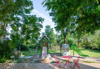 Speelplein en terras onder de bomen bij Camping Vauban, een vakantiepark in Grand Est, Frankrijk.