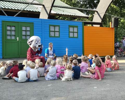 Kinder sitzen vor einer Person im Hundekostüm und einem Erwachsenen bei einer Aufführung auf einem Campingplatz.