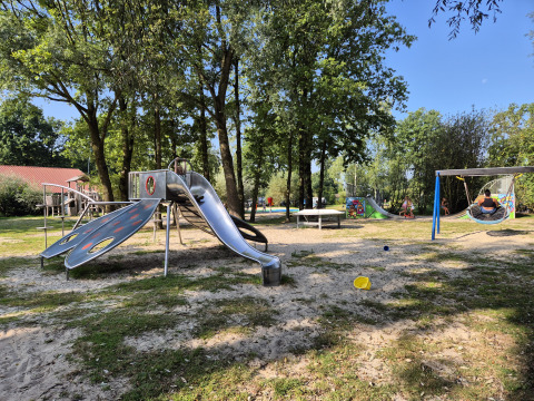 Spielplatz auf dem Campingplatz De Rammelbeek in Overijssel, Niederlande, mit Rutsche, Schaukel und Bäumen.