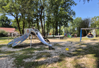 Spielplatz auf dem Campingplatz De Rammelbeek in Overijssel, Niederlande, mit Rutsche, Schaukel und Bäumen.