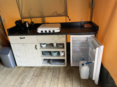 Compact kitchen with black countertop, stove, open fridge, and dishes inside a safari tent interior.