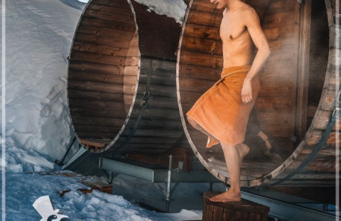 Uomo esce da una sauna tonda in legno con asciugamano, circondato dalla neve a Iglo Lodge Burgberg.