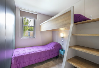 Room with a bunk bed, single bed, purple bedding, window, and wooden stairs at Mobile home Confort in France.