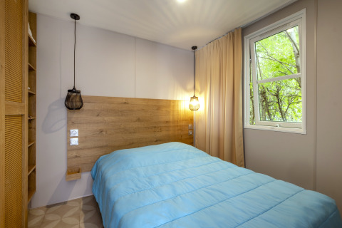 Modernes Schlafzimmer im Mobile Home Confort, Seasonova Camping Le Plan d’Eau, mit blauem Bett und Fenster.