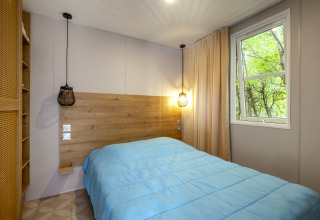 Modernes Schlafzimmer im Mobile Home Confort, Seasonova Camping Le Plan d’Eau, mit blauem Bett und Fenster.