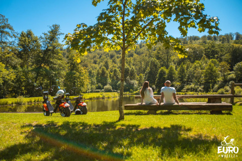 Paar entspannt an einem See bei Glamping, mit zwei Elektrorollern und Bäumen im Hintergrund.