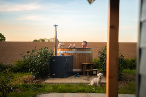 Pareja en jacuzzi en Berg Lodge Tiny House, paisaje rural, perro pequeño sentado en primer plano.