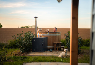 Pareja en jacuzzi en Berg Lodge Tiny House, paisaje rural, perro pequeño sentado en primer plano.