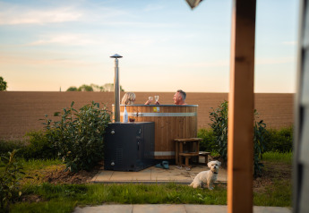 Couple relaxing in hot tub at Berg Lodge Tiny House, rural setting, small dog sitting in foreground.