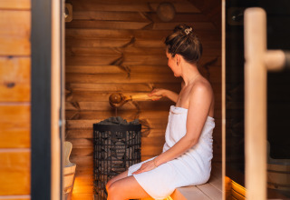 Vrouw met handdoek zit in sauna en giet water op hete stenen, wellness bij glamping accommodatie.