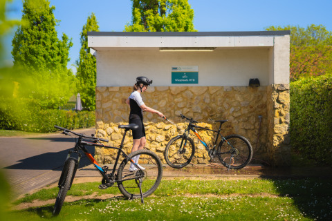 Une personne lave un VTT à une station de lavage extérieure dans un hébergement glamping entouré de verdure.