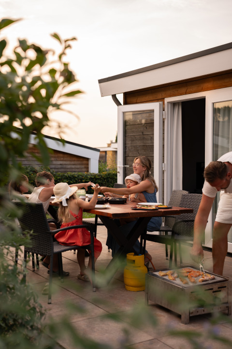 En familie nyder udendørs middag med grill foran et moderne glampinghus i aftensolen.