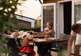 En familie nyder udendørs middag med grill foran et moderne glampinghus i aftensolen.