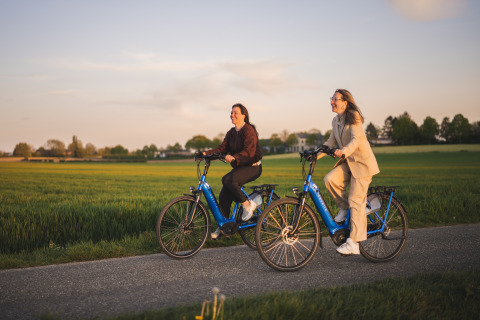 Zwei Frauen fahren auf blauen E-Bikes bei Sonnenuntergang an einer Glamping-Unterkunft durch grüne Felder.