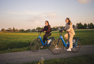 Due donne pedalano su biciclette elettriche blu accanto a una sistemazione glamping al tramonto in campagna.
