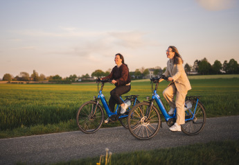 To kvinder cykler på blå elcykler gennem det grønne landskab ved en glampingindkvartering i solnedgang.