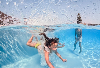 Børn leger og svømmer under vandet i en swimmingpool på glamping, med vandstænk over overfladen.