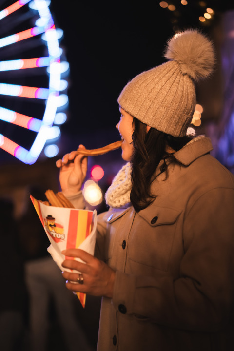 Frau genießt Churros bei einer Glamping-Unterkunft am Abend, Riesenrad und Lichter im Hintergrund.